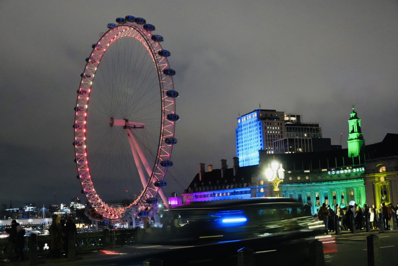 London Eye lit pink at night with a black cab passing in the foreground, motion blurred