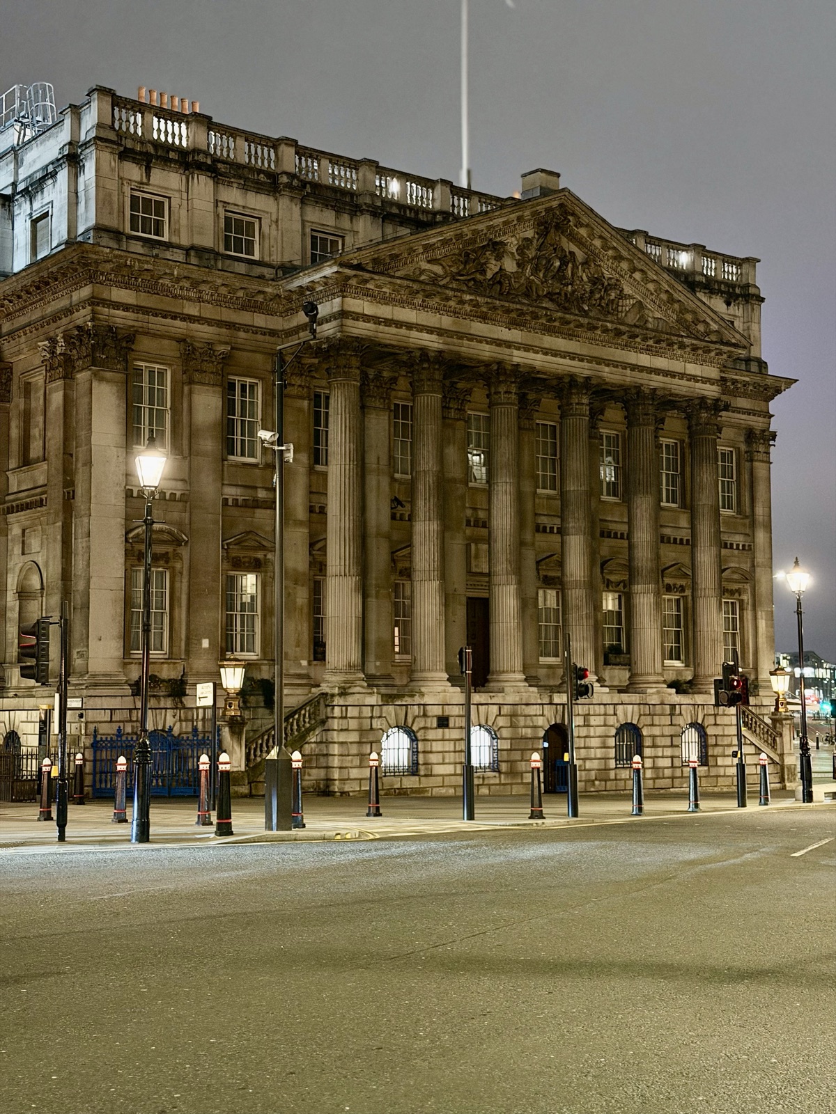 A grand neoclassical building with columns, lit warmly at night, empty street in front